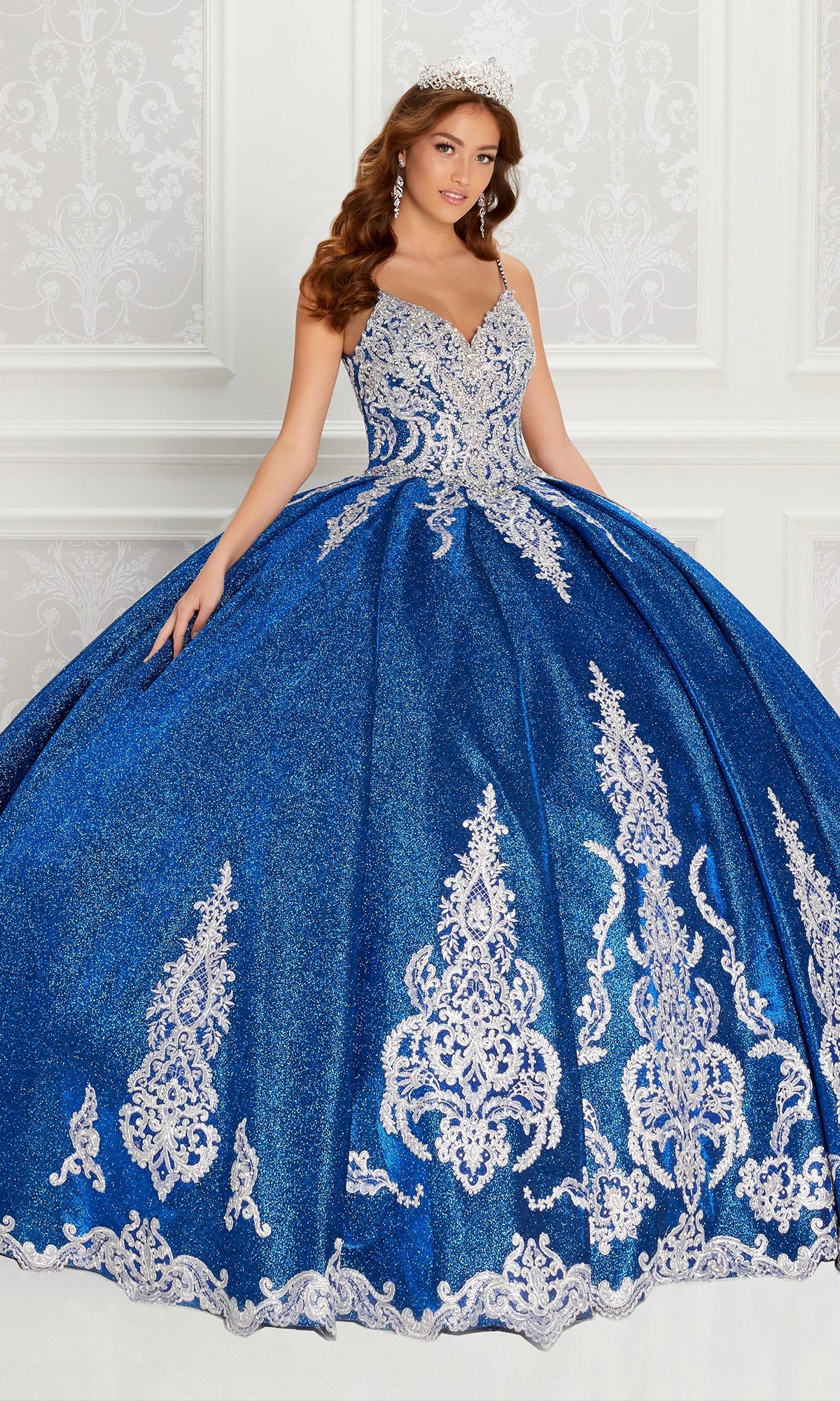 A woman in the Quinceanera Dress PR22141 by Princesa, a sparkling royal blue ball gown with silver embroidery and corset back, wears a tiara as she stands before an ornate white background.
